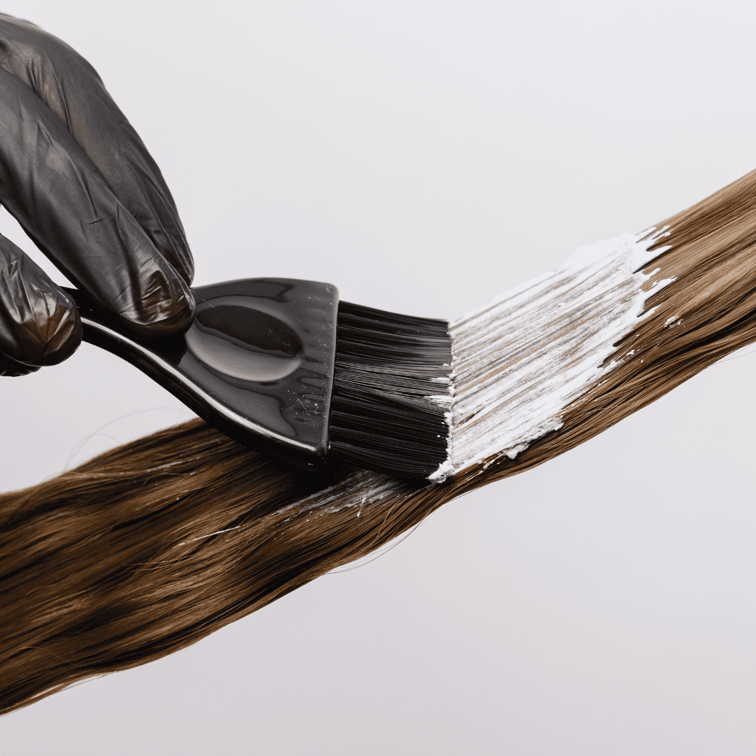 Applying hair dye with a brush onto brown hair strands wearing black gloves.